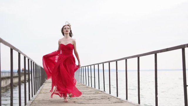 Pretty girl is wearing silk red dress is running on a wooden pier. She is holding her swaying skirt and looking forward, romantic and fashion shot