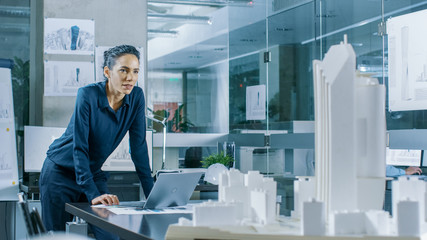 Female Architectural Designer Works on a Laptop,  Engineering New Building Model for the Urban Planning Project. Clean Minimalistic Office, Concrete Walls Covered by Blueprints and Documents.