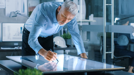 Senior Architectural Engineer Draws Building Concept on a Graphics Tablet Display Vertical Touchscreen Table. Clean Minimalistic Office, Concrete Walls Covered by Blueprints.