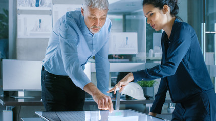 Senior Male and Young Female Architectural Designers Draw Building Concept on a GraphicsTablet Display Vertical Touchscreen Table. They Use Gestures for Zooming Project Model.