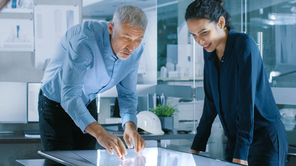 Senior Male and Young Female Architectural Designers Draw Building Concept on a Graphics Tablet Display Vertical Touchscreen Table. They Use Gestures for Zooming Project Model.