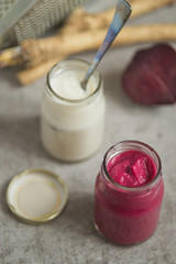 Two small glass jars with seasoning, fresh horseradish root, beets and a metal grater on a gray background. 