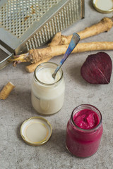 Two small glass jars with seasoning, fresh horseradish root, beets and a metal grater on a gray background.