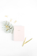 Pale pastel pink notebook, golden pen and clips, monstera palm leaf decoration on white background. Flat lay, top view home office desk concept.