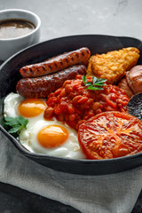 Full English breakfast with bacon, sausage, fried egg, baked beans, hash browns and mushrooms in rustic skillet, pan. cup coffee.