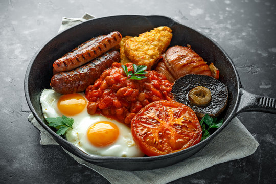 Full English breakfast with bacon, sausage, fried egg, baked beans, hash browns and mushrooms in rustic skillet, pan.