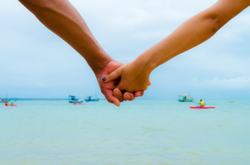 Couple holding hands having the sea as a background. View of the arms in the center. Some boats in blur.