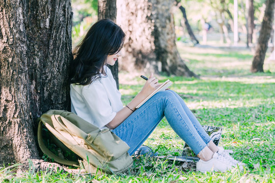 Woman relaxing with pen writing on a notebook sitting under a tree on grass in park