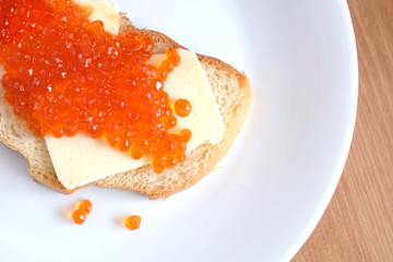 Sandwich with butter and red salmon caviar on white bread lies on white round plate on wooden table top view