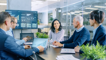 Chief Female Executive Gives a Report to Her Colleagues in the Meeting Room. Diverse Team of Smart People Listens Carefully. Modern Looking Office Has Wall TV with Statistics Shown on it.