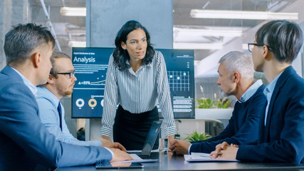Chief Female Executive Leans and Spreads Project Blueprints on the Table Showing them to Her Colleagues. In the Background Pie Charts and Company's Growth on the Wall TV.