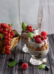 Two glasses of chia pudding with fresh strawberries, raspberries and blueberries. Basket with berries. On a wooden grey background.
