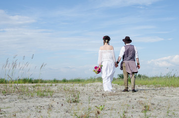 Boho wedding couple. Back view. Stylish hippie woman in white ethnic dress holding hands with man