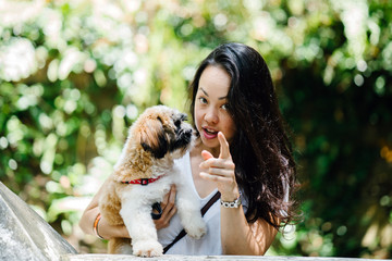 Image of a young Pan Asian woman with her favorite dog and smiling for the camera in a green park on a warm day in the park. The dog is a toy breed shih tzuh.