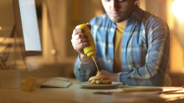 Cinemagraph of young man sitting at table with computer and squeezing extra mustard on appetizing burger