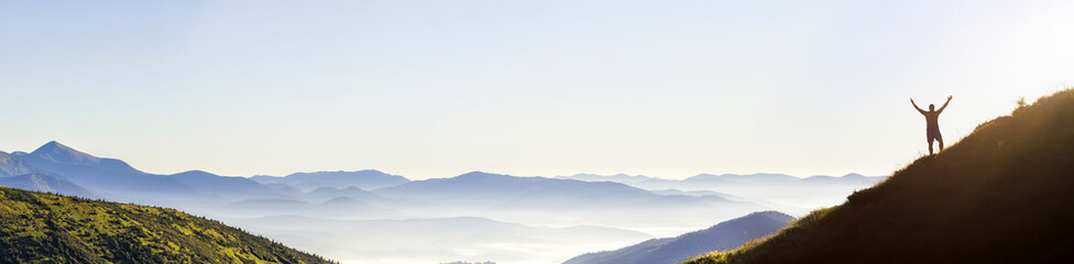 Panorama of young successful man hiker silhouette open arms on mountain peak.