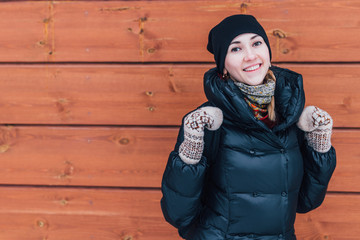 Young smiling woman in winter cloths standing in front of wooden wall outdoors