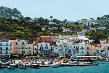 Fototapeta na wymiar Italy stunning view of Capri when arriving to the island by ferry. Numerous boats and ferries at the dock. A lot of peole on the beach