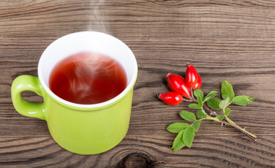Rosehip tea and red fruits of wild rose on brown wood background. Rosa canina. Green mug with smoking medicinal drink and decorative twig with rosehips, leaves and thorns.