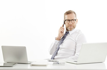 Busy male broker with mobile phone. Middle aged happy businessman using mobile phone and making call while sitting at desk in front of laptop. Isolated on white background.