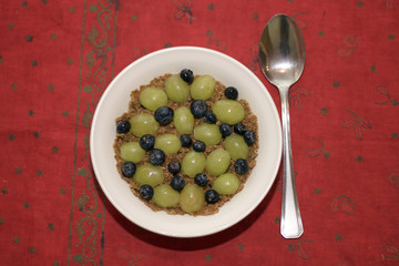 A bowl of cereal with sliced grapes and whole blueberries on top displayed on a Christmastime table cloth.