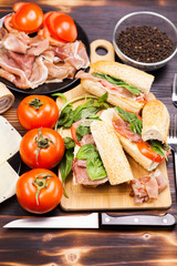 Three healthy and delicious sandwiches on wooden board next to tomatoes, greenery, ham and cheese