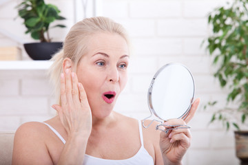 Middle aged woman looking at wrinkles in mirror. Plastic surgery and collagen injections. Makeup....