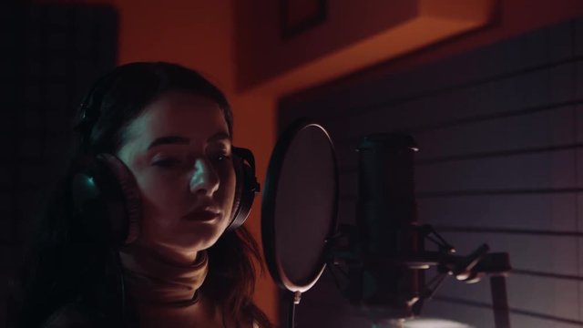 Beautiful young white girl wearing headphones and singing into microphone in recording studio