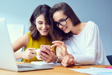 Couple of cheerful businesswomen browsing on phone and enjoying coffee in the office