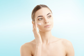 Studio shot of beautiful young woman applying moisturizer cream onto her face against at isolated light blue background with copy space. 