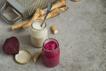 Two small glass jars with seasoning, fresh horseradish root, beets and a metal grater on a gray background. 