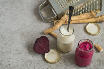 Two small glass jars with seasoning, fresh horseradish root, beets and a metal grater on a gray background. 