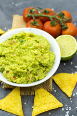Avocado dip guacamole with tortilla chips in a white bowl on a black stone table. Traditional Mexican appetizer.