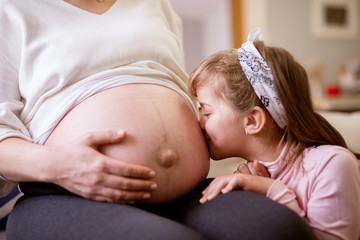 Adorable little toddler girl kissing belly of her pregnant mother. Family and new life concept.