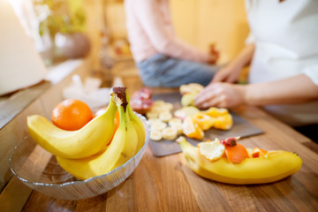 Close up focus view of bananas and oranges while pregnant woman preparing fruit salad while her pretty toddler daughter sitting on the kitchen table next to her.