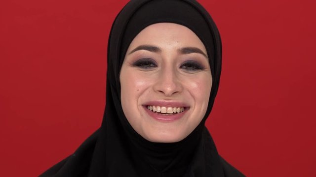 Portrait of middle eastern woman with smoky eyes visage putting off black niqab or burqa and smiling on camera, over red background. Concept of emotions