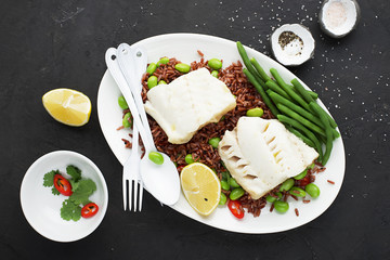 Juicy fresh steam white sea fish fillets on a cushion of red fragrant rice and fresh vegetables: soybeans, capsicum, lemon. Top View.
