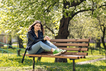 Glamorous young Caucasian woman in black leather jacket sitting in the park on the bench