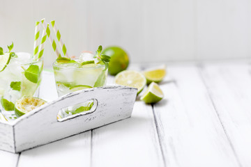 Caipirinha, mojito cocktail with lime, brown sugar, ice and mint leaves in beautiful glasses, cut green citrus on white wooden background. Summer alcohol drink. Close up photography. Selective focus