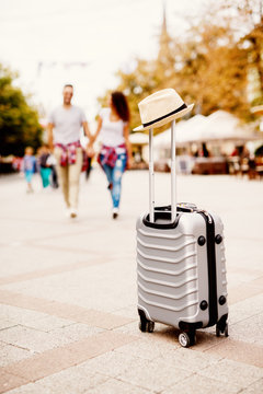 Photo of travelling bag with a hat and couple walking while holding hands in the background.