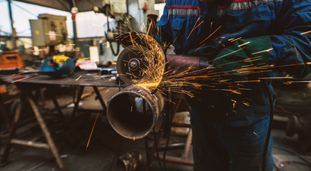 Fabric worker in protective uniform cutting metal pipe on the work table with an electric grinder in the industrial workshop.