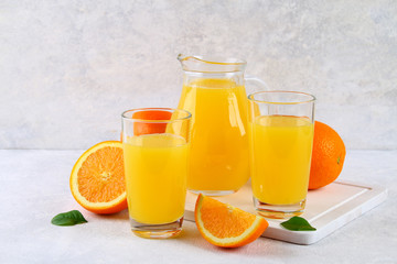 Glass cups and a pitcher of fresh orange juice with slices of orange and yellow tubes on a light gray table.