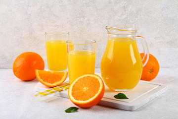 Obraz na płótnie Canvas Glass cups and a pitcher of fresh orange juice with slices of orange and yellow tubes on a light gray table.