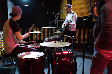 Obraz na płótnie Canvas Band of young musicians performing in dim recording studio making new album, drum set in foreground