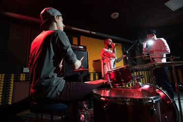 Portrait of modern music band rehearsing in recording studio lit by dim lights while making new album, drummer in foreground