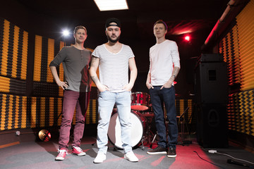 Full length portrait of modern music band posing in recording studio and looking at camera with...
