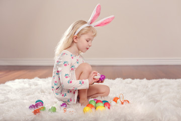 Cute adorable Caucasian child girl wearing Easter bunny rabbit ears sitting on soft fluffy rug carpet in studio. Kid holding holiday colorful eggs celebrating traditional holy Christian holiday