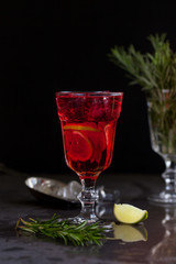 a glass with a cocktail of red color with lime, rosemary and ice cubes on a background of rosemary on a dark background