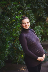 Portrait of a smiling beautiful pregnant woman with a sparkling look in a knitted sweater holding on to her belly in the park. A cute girl is standing in the garden on a background of green foliage.