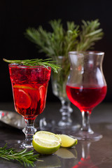 a glass with a cocktail of red color with lime, rosemary and ice cubes on a background of rosemary on a dark background